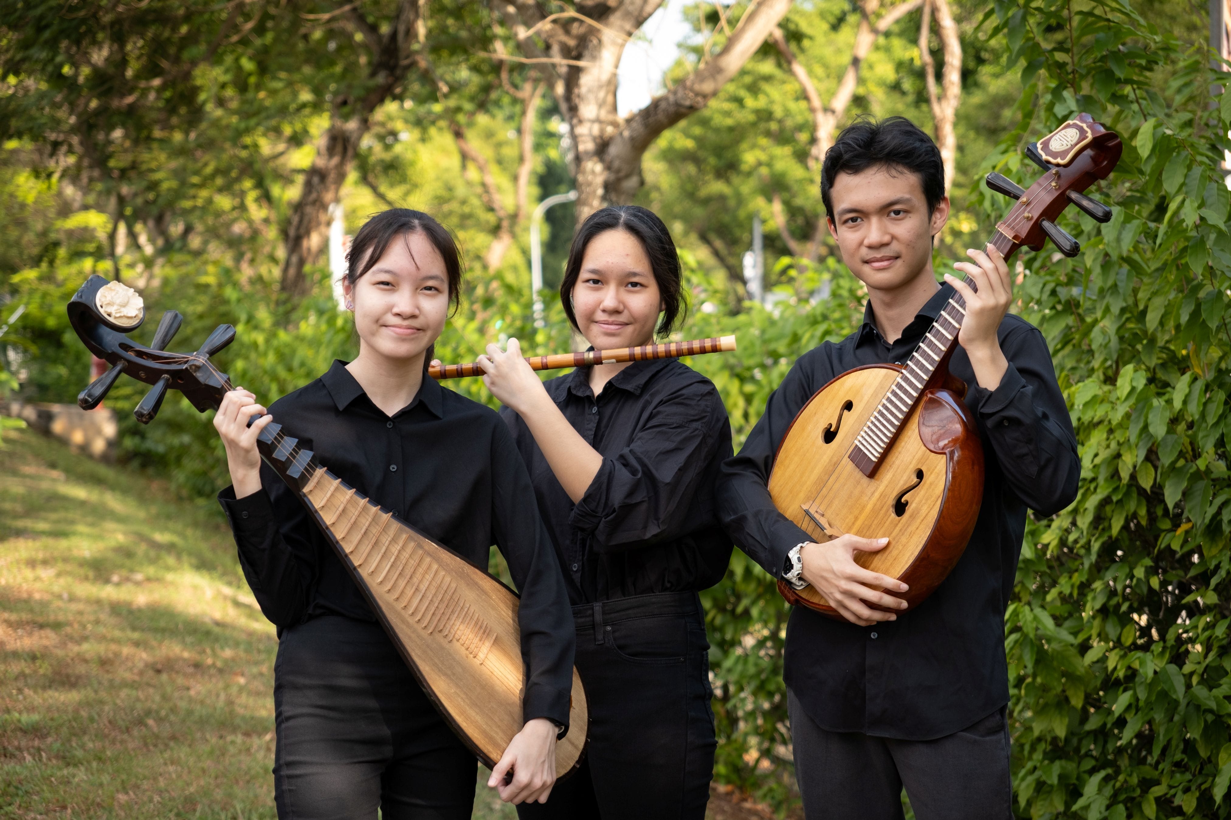 Three students in black attire pose outdoors against a lush green backdrop. The student on the left holds a pipa (a pear-shaped lute), the student in the centre rests a dizi (a bamboo flute) on her shoulder, and the student on the right holds a zhongruan (a larger round-bodied lute).